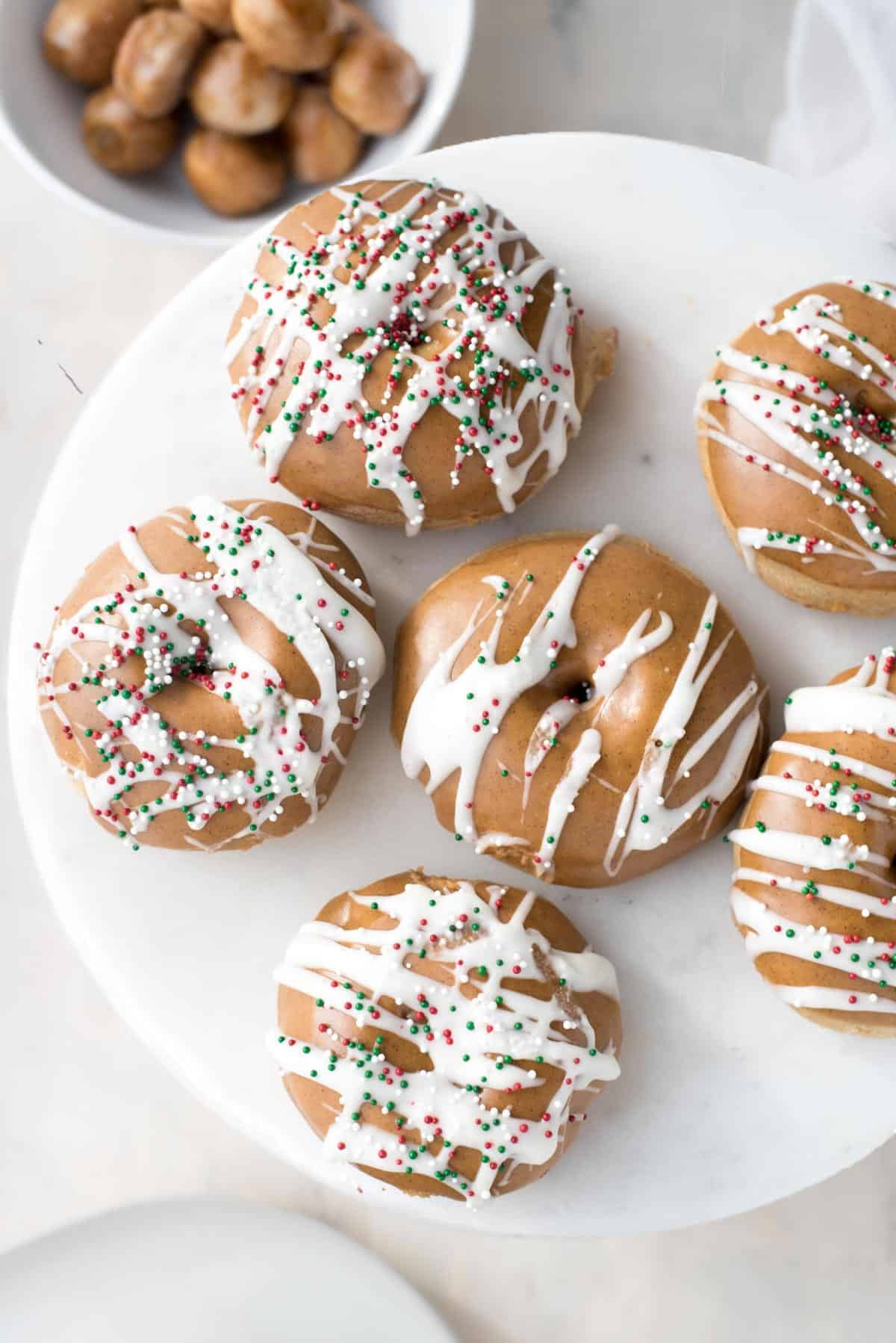 View form above of fluffy baked gingerbread doughnuts. 5 doughnuts in a ring with a 6th in the middle on a large white plate. Each doughnut is topped with a brown colored gingerbread glaze and white icing has been drizzled decoratively on top, along with festive colored sprinkles. To the top left of the doughnuts is a white bowl filled with doughnut holes.