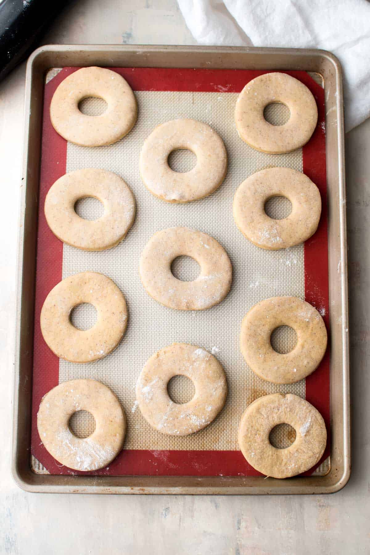 The doughnut dough has been cut into the O shape with a hole in the middle. They are sitting on a silver baking tray with a red rim. At this point they dough is quite flat.