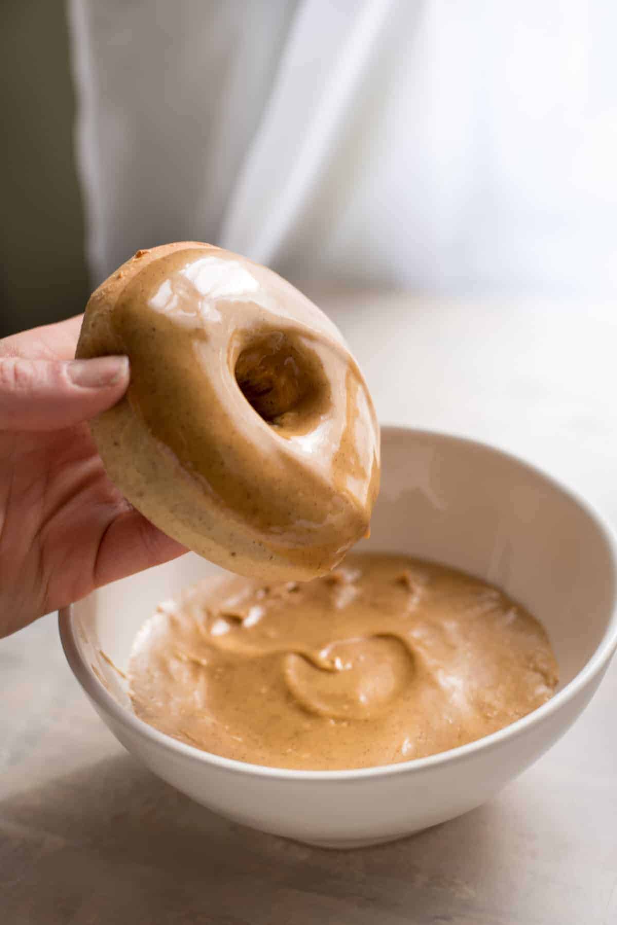 A white bowl contains gingerbread glaze. A hand is holding a doughnut which has been dipped into the glaze on top. The hand is holding the doughnut over the bowl to catch ay excess drips.