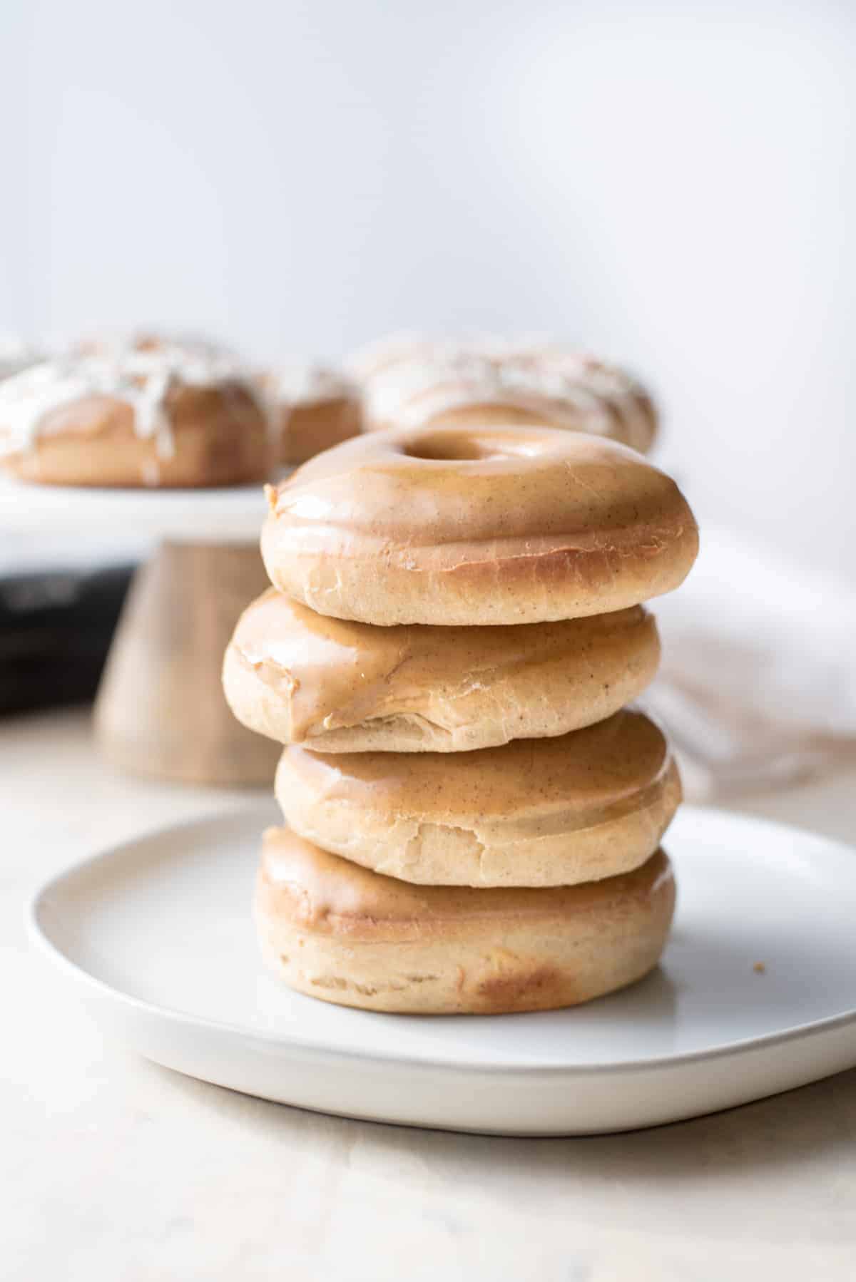 4 baked gingerbread doughnuts sitting on top of each other in a column, sitting on a white ceramic plate. In the background there is a raised cake dish displaying some more doughnuts, they are out of focus.