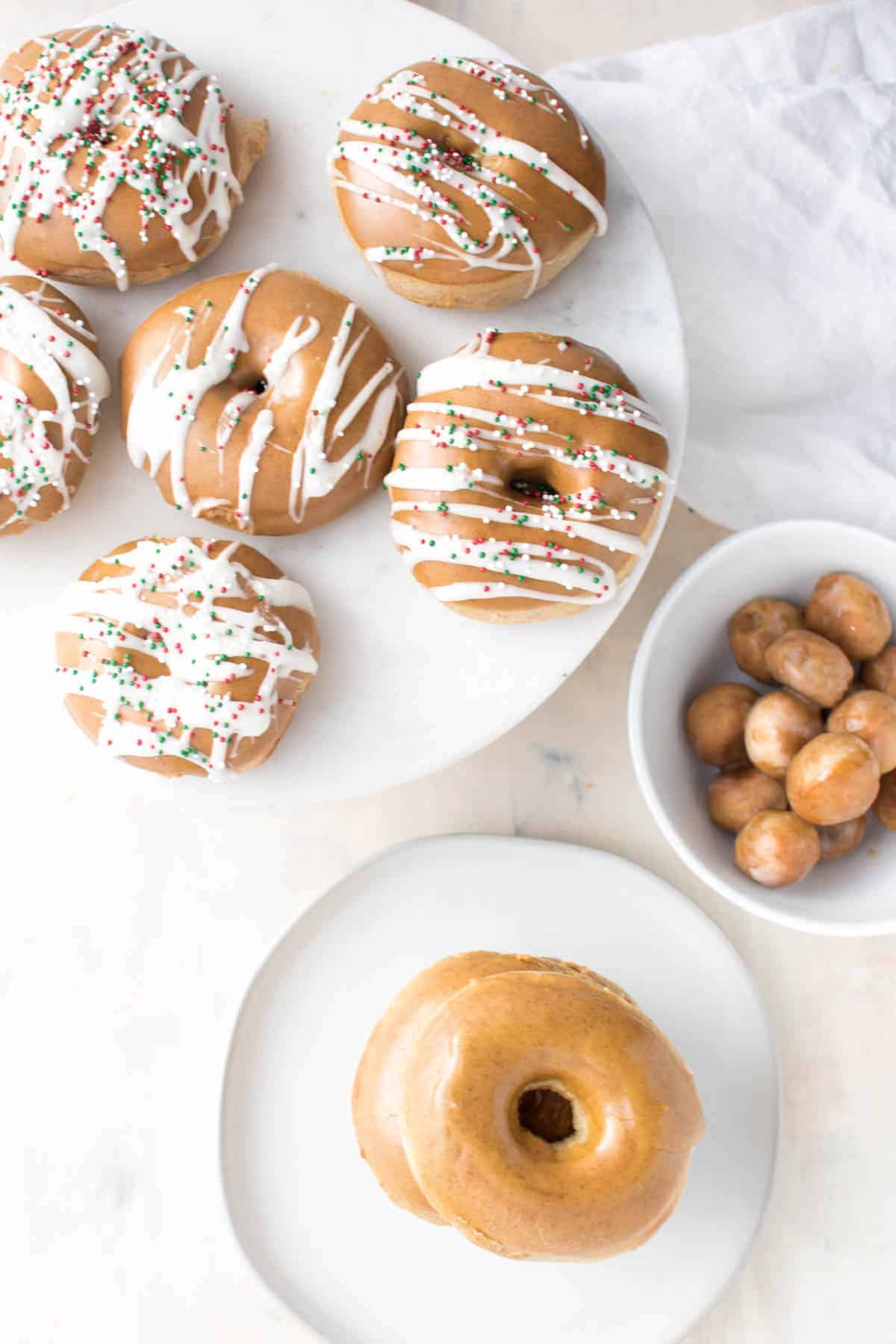 Glazed Gingerbread Doughnuts sitting in a circle on a white ceramic plate. There are 6 doughnuts, 5 in a circle, 1 in the middle. They have been decorated with a white colored frosting in lines from side to side across the doughnut and a few red and green sprinkles. Next to the plate is a small white bowl filled with glazed doughnut holes that have been cut out from the doughnuts. There is a final small white plate with 2 glazed doughnuts, sitting directly on top of each other with no decoration.