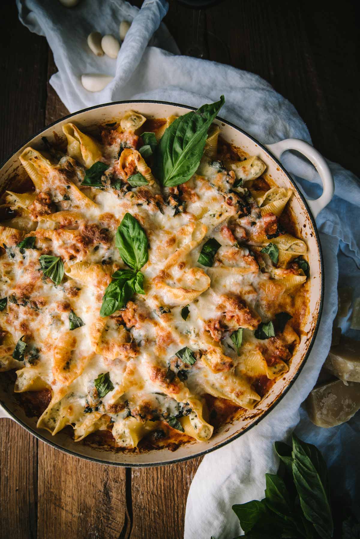 View from above of baked ricotta stuffed shells in a large white braiser with handles. The edges are crisp and there is lota of melted cheese that has bubbled on the top. Fresh basil leave are used to garnish the top.