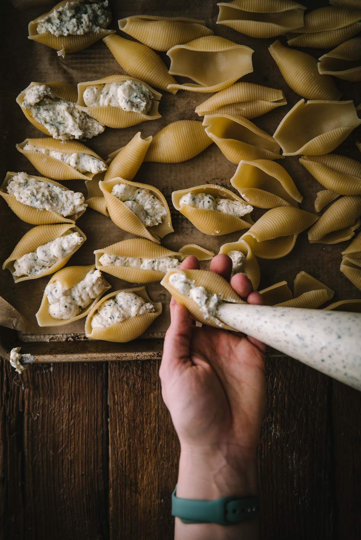 Someone is piping ricotta mixture into jumbo shell pasta through a piping bag. They are holding one shell in their hand while they pipe. A lined sheet try lays underneath where all the other stuffed shells are resting.