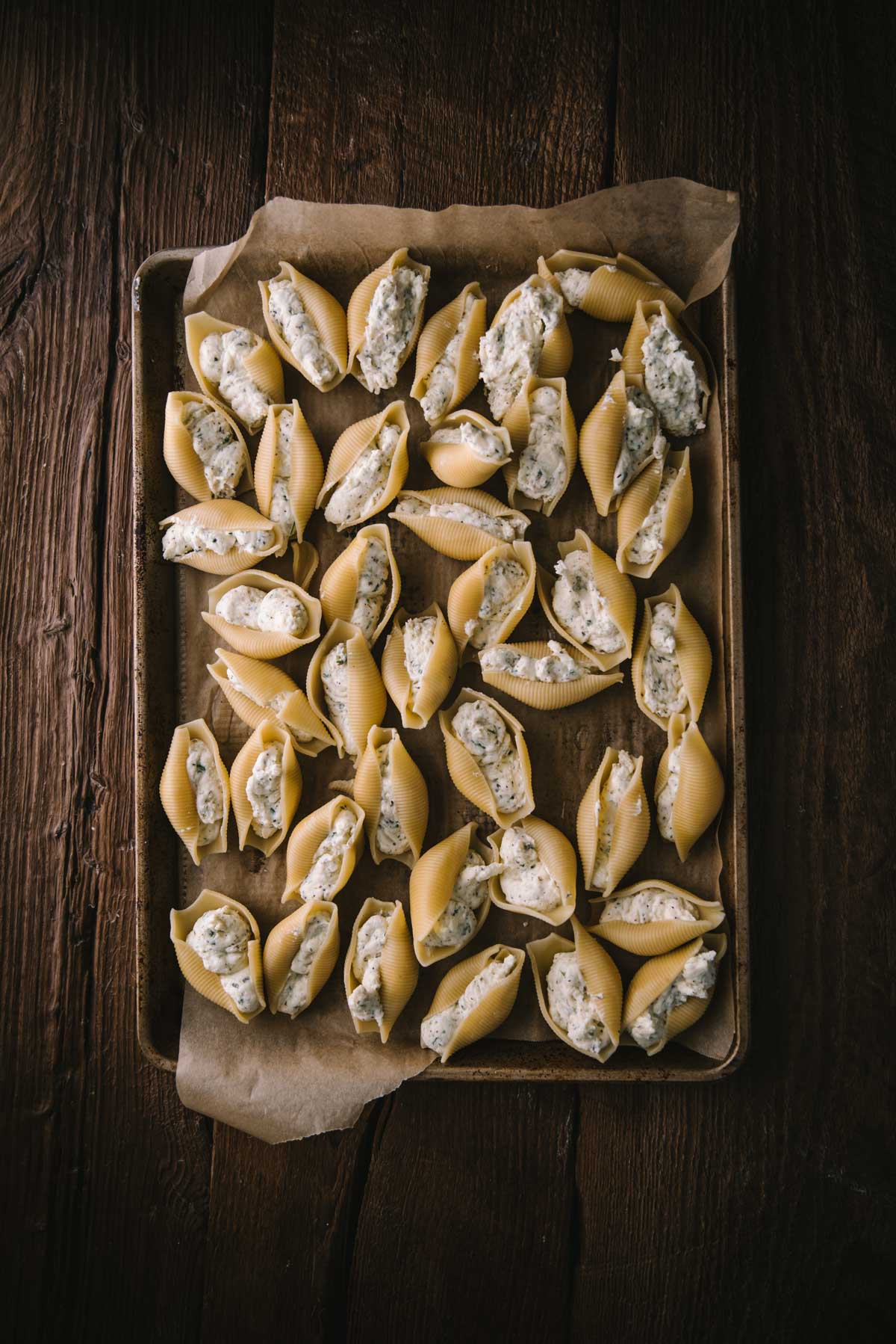 Pasta shells have been stuffed with the ricotta mixture and are sitting on a sheet tray on a wooden table, ready to be assembled.