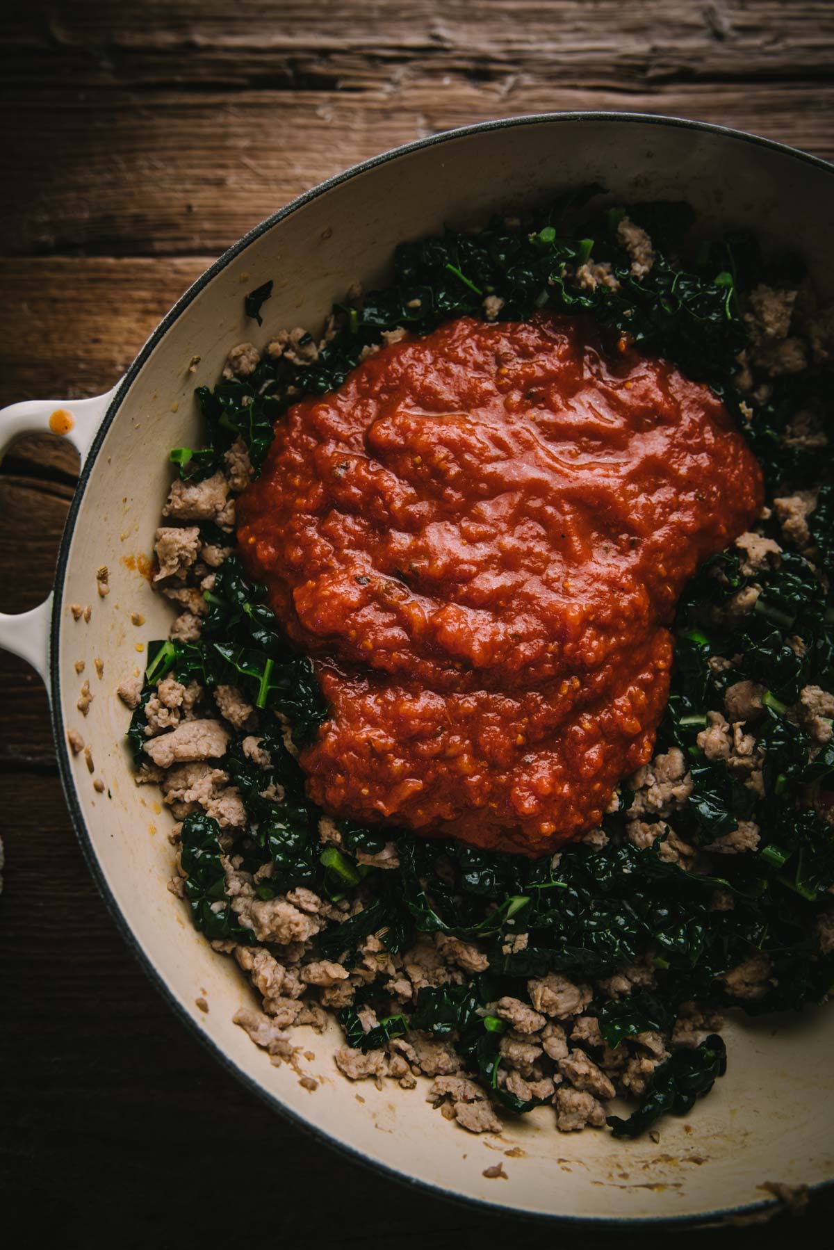 Sausage meat and kale marinara sauce are in a white braiser pan with handles. The ingredients have not yet been combined.