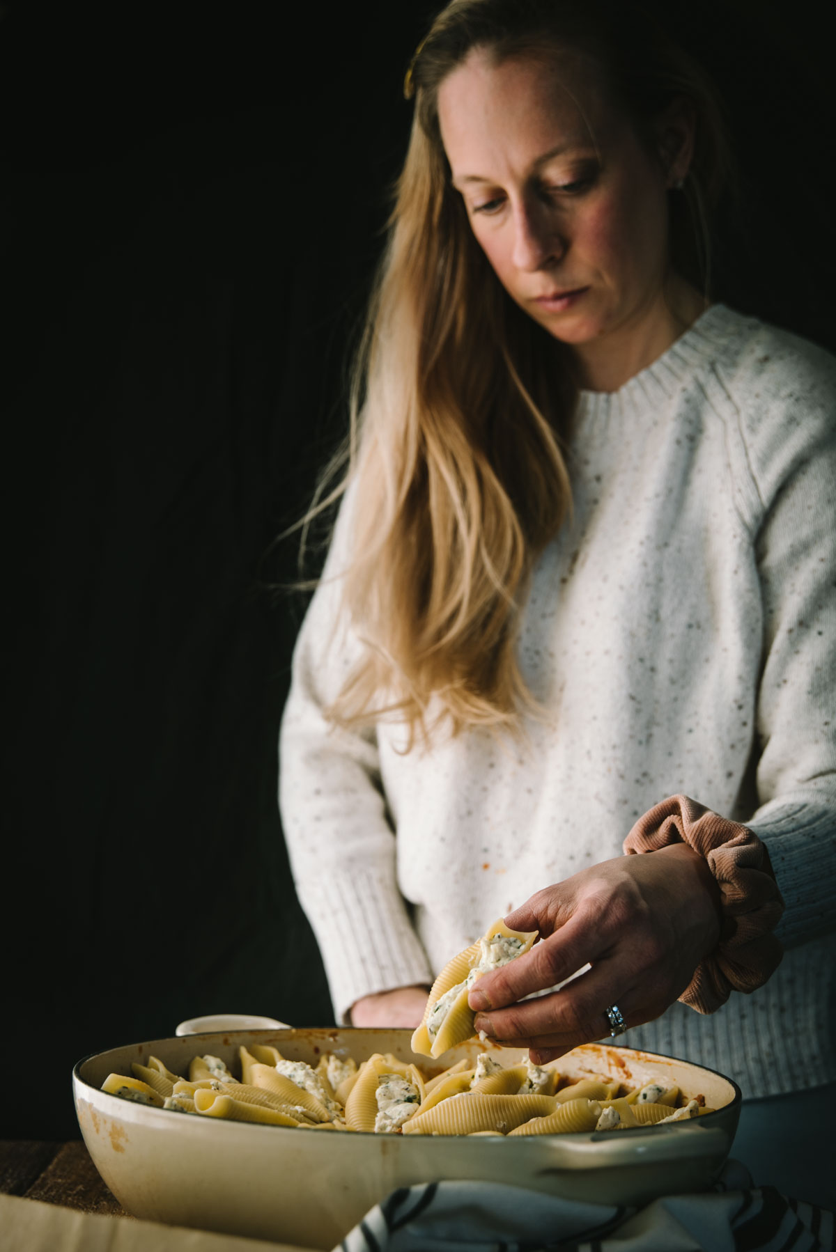 Someone wearing a white jumper is standing in a dark room over a white oven safe pan and is placing jumbo shells in the pan for baking. They are about to lay a shell down in this photo.