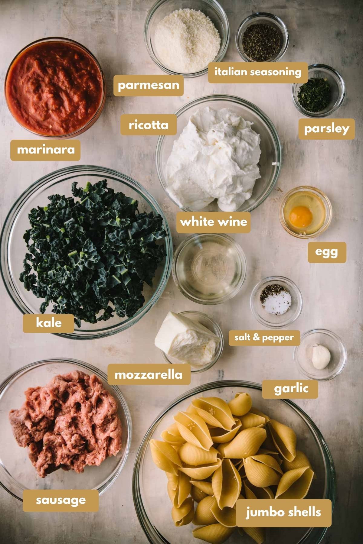 All the ingredients required to make ricotta stuffed shells in clear glass bowls with labels. There is marinara sauce, parmesan cheese, Italian seasoning, parsley, ricotta, an egg, white wine, kale, salt and pepper, mozzarella, garlic, sausage meat and jumbo pasta shells