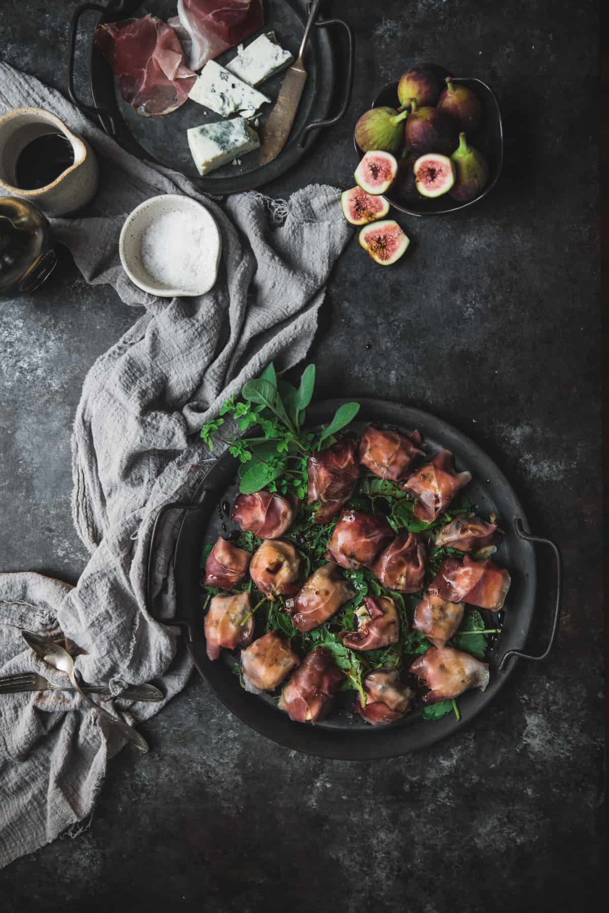 overhead angle of prosciutto wrapped figs on a silver tray with extra ingredients around it