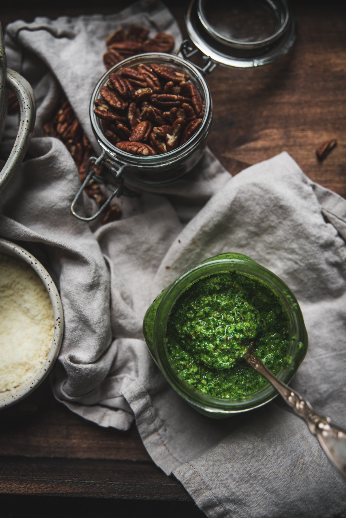 spinach pecan pesto in a glass jar with a spoon and ingredients in other jars