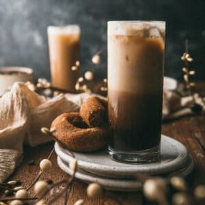 Layers of shaken espresso, ice, and milk in a glass on a plate with cider donuts