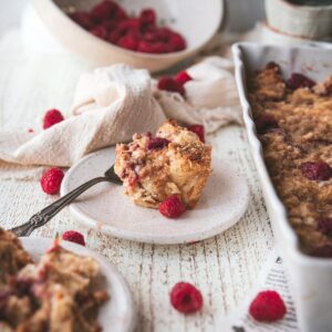 piece of raspberry cheesecake french toast bake on a plate with a fork surrounded with fresh berries