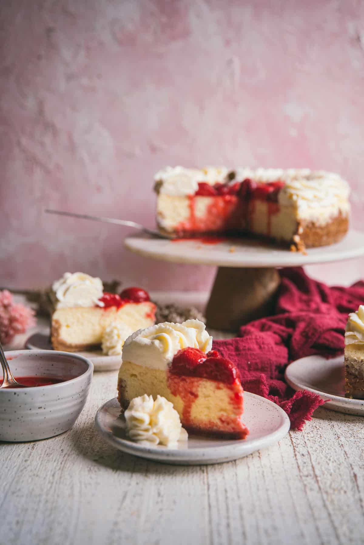 three slices of cheesecake on plates with whipped cream garnish with a strawberry topped cheesecake on a cake stand in the background