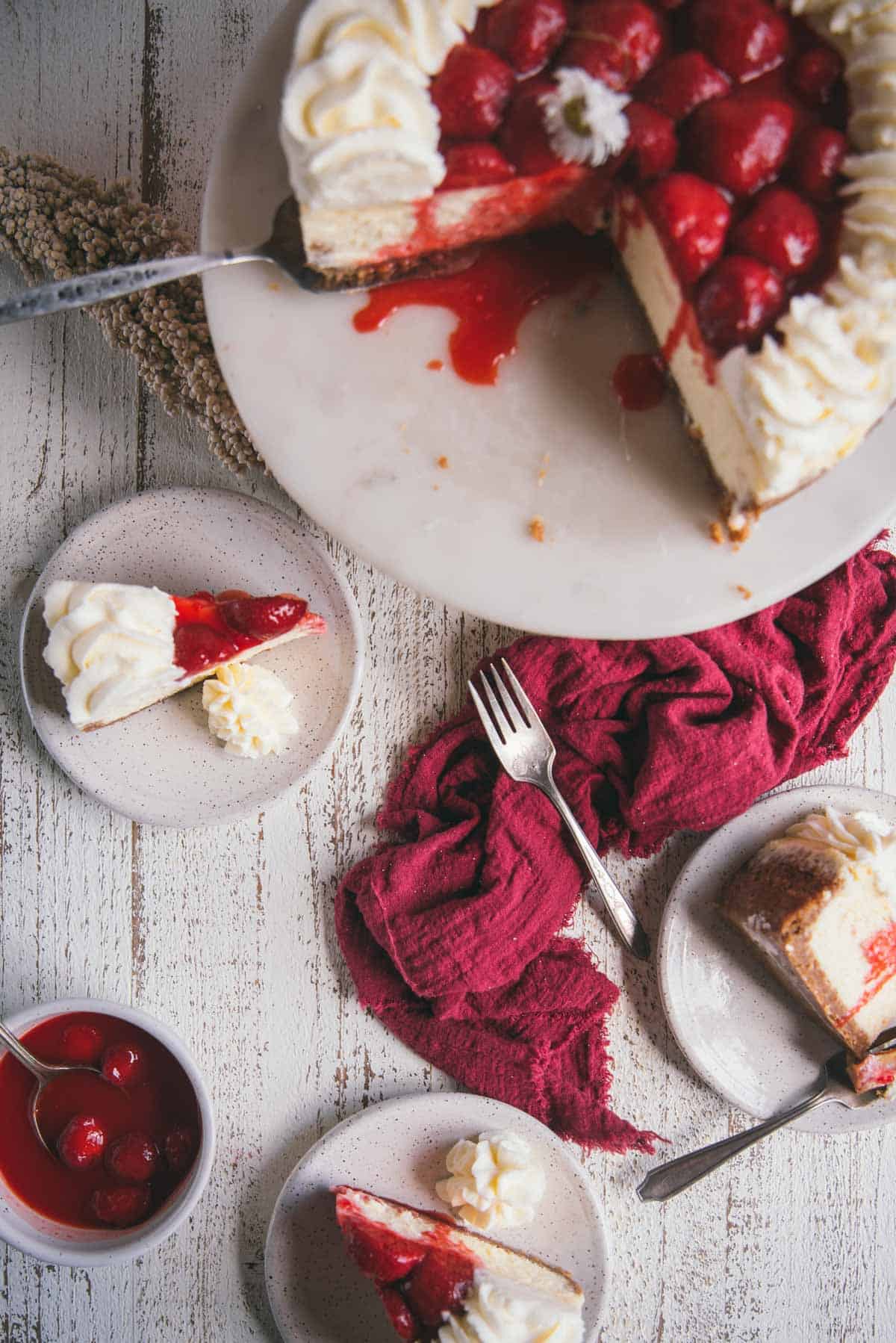 overhead image of slices of cheesecake on top of plates with a fresh strawberry cheesecake on a cake stand out of focus