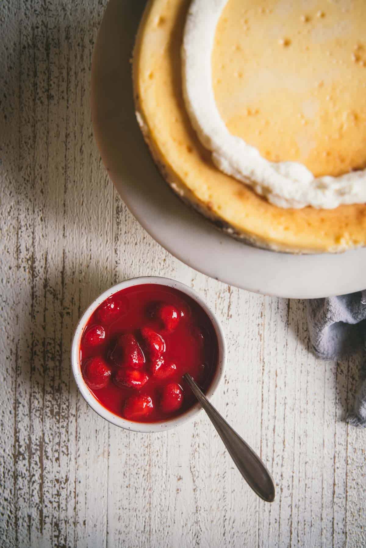 circle of whipped cream on top of a cake next to a bowl of strawberry sauce with a spoon in it