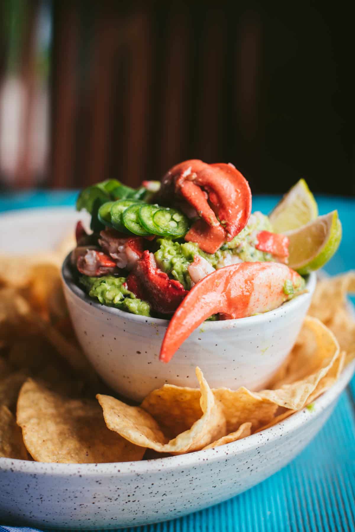 Lobster guacamole in a white terrazzo bowl. The lobster tails are large and lay slightly over the edge of the bowl, there is fresh jalapenos on top and 2 wedges of lime.
The bowl sits on a white terrazzo plate filled with chips to dip.