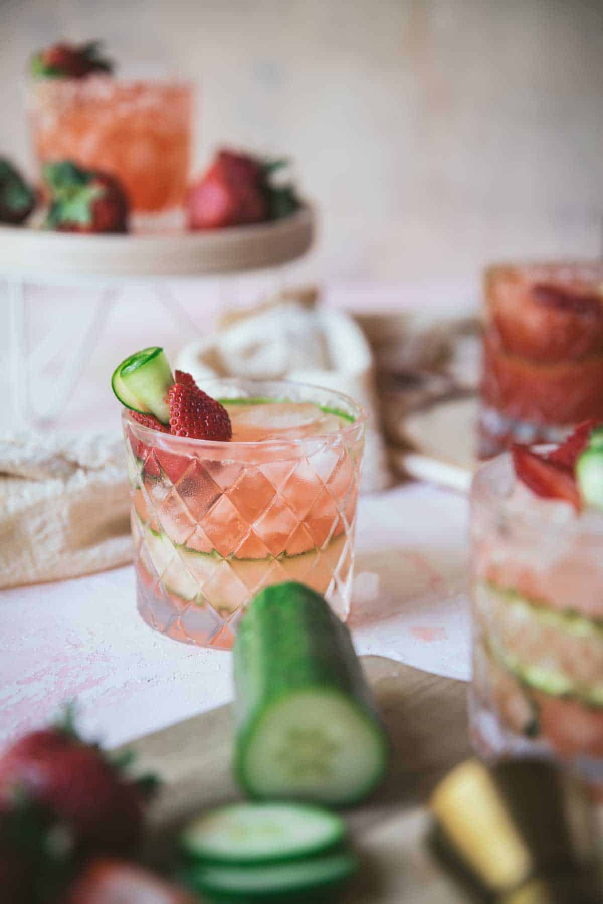 Completed Strawberry Cucumber Margarita in decorative rocks drinking glass, poured over ice, garnished with a cucumber scroll and cut strawberries. Sitting on a white tablecloth.
Out of focus, there are 3 other (partial view) completed cocktails and leftover garnish of cucumber on a wooden chopping board and strawberries. 