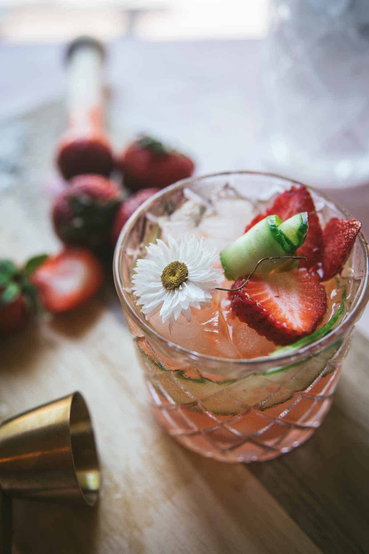 Close up image of completed cocktail. Pink cocktail in decorative rocks glass. A cucumber peel is wrapped around the inside of the glass, it is granished on top with sliced strawberries, cucumber scrolls and a daisy. There are additional chopped strawberries in the background and a brass measuring cup.