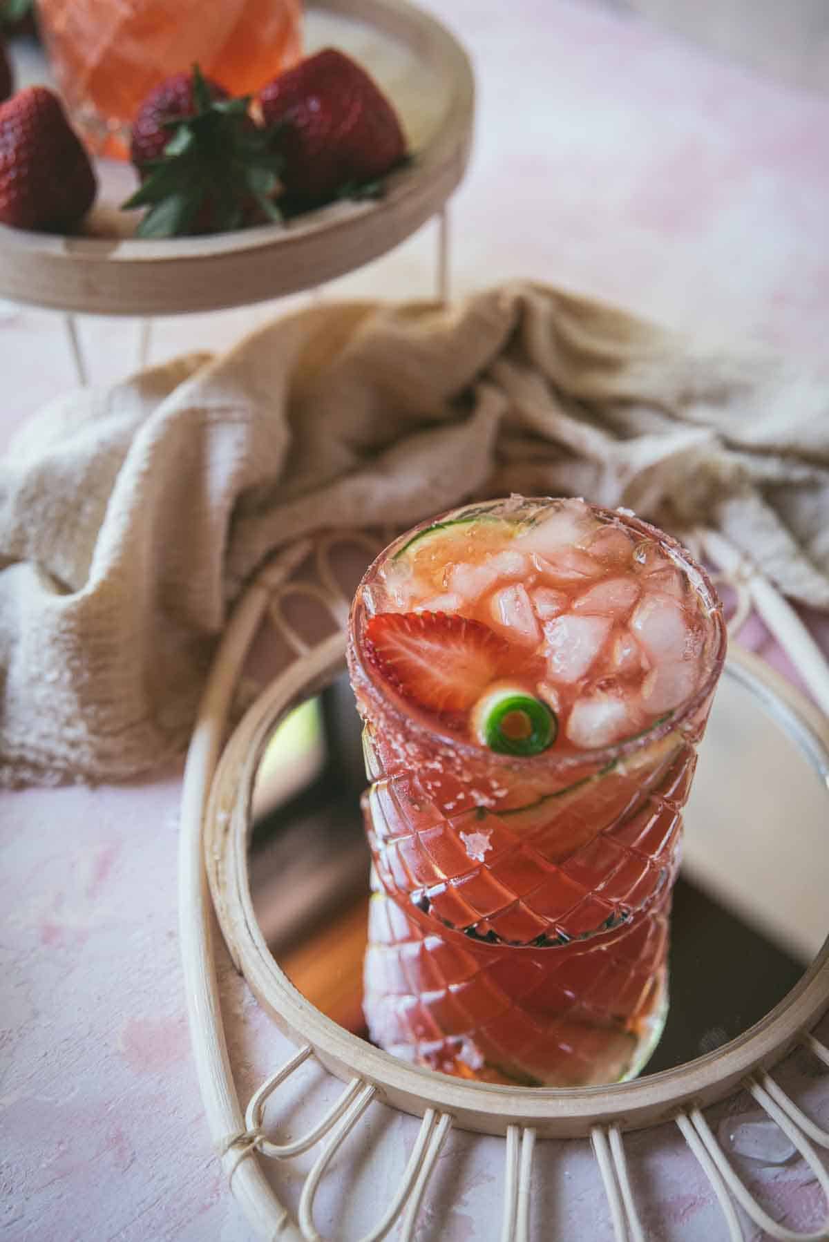 Completed pink cocktail, in decorative rocks glass. Topped with lots of ice, cucumber scroll and a strawberry slice. The drink sits on top of a boho style wicker mirror. A cloth is nearby and a short table with additional strawberries and another cocktail.