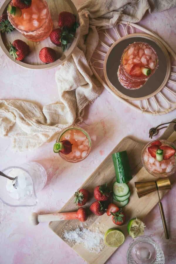 overhead of ingredients and completed strawberry cucumber margaritas with linen napkin on pink background