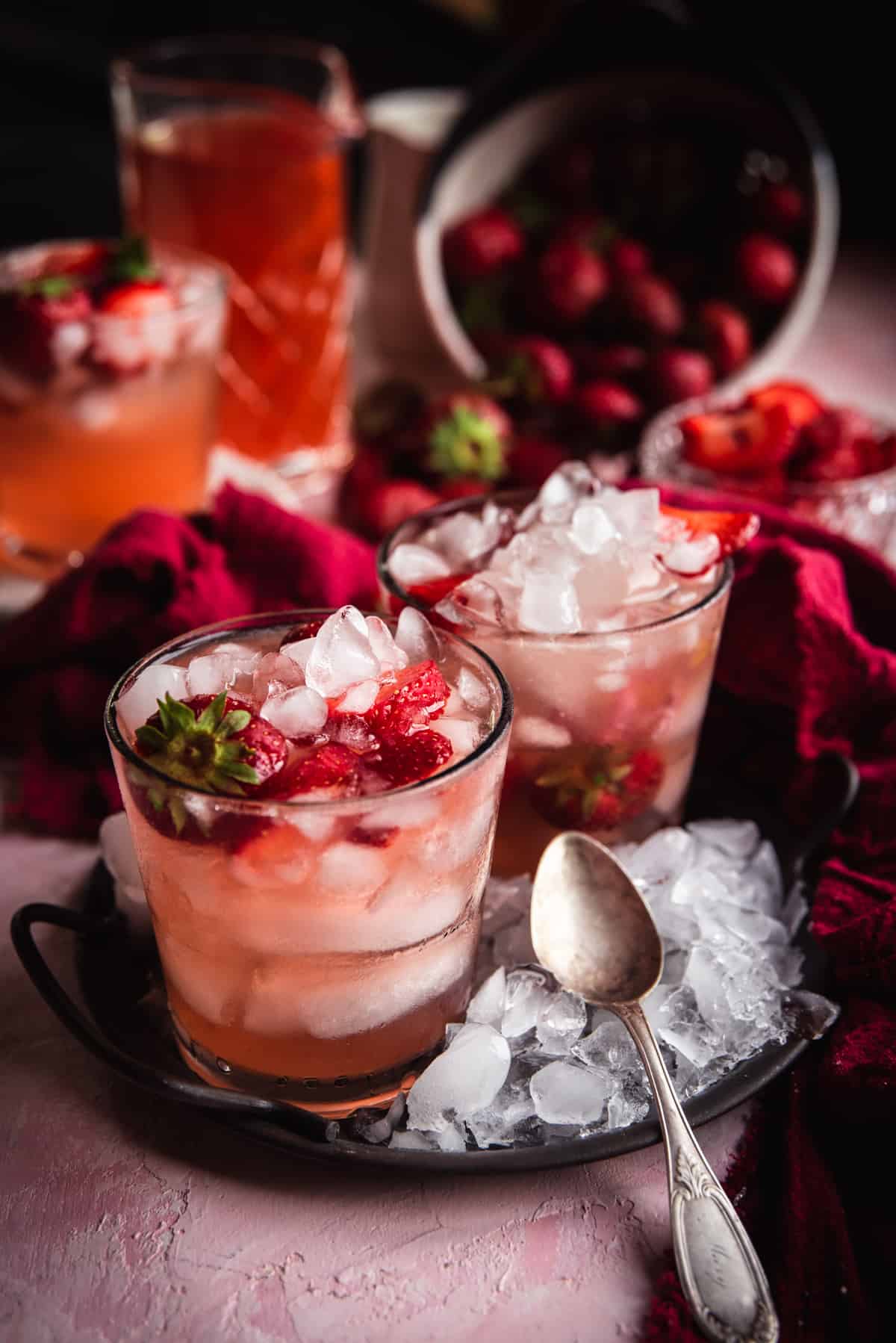 two glasses with ice and pink sangria on a circlular metal serving dish covered in crushed ice and vintage spoon on top.  Pitcher of sangria and bowl of strawberries in the background