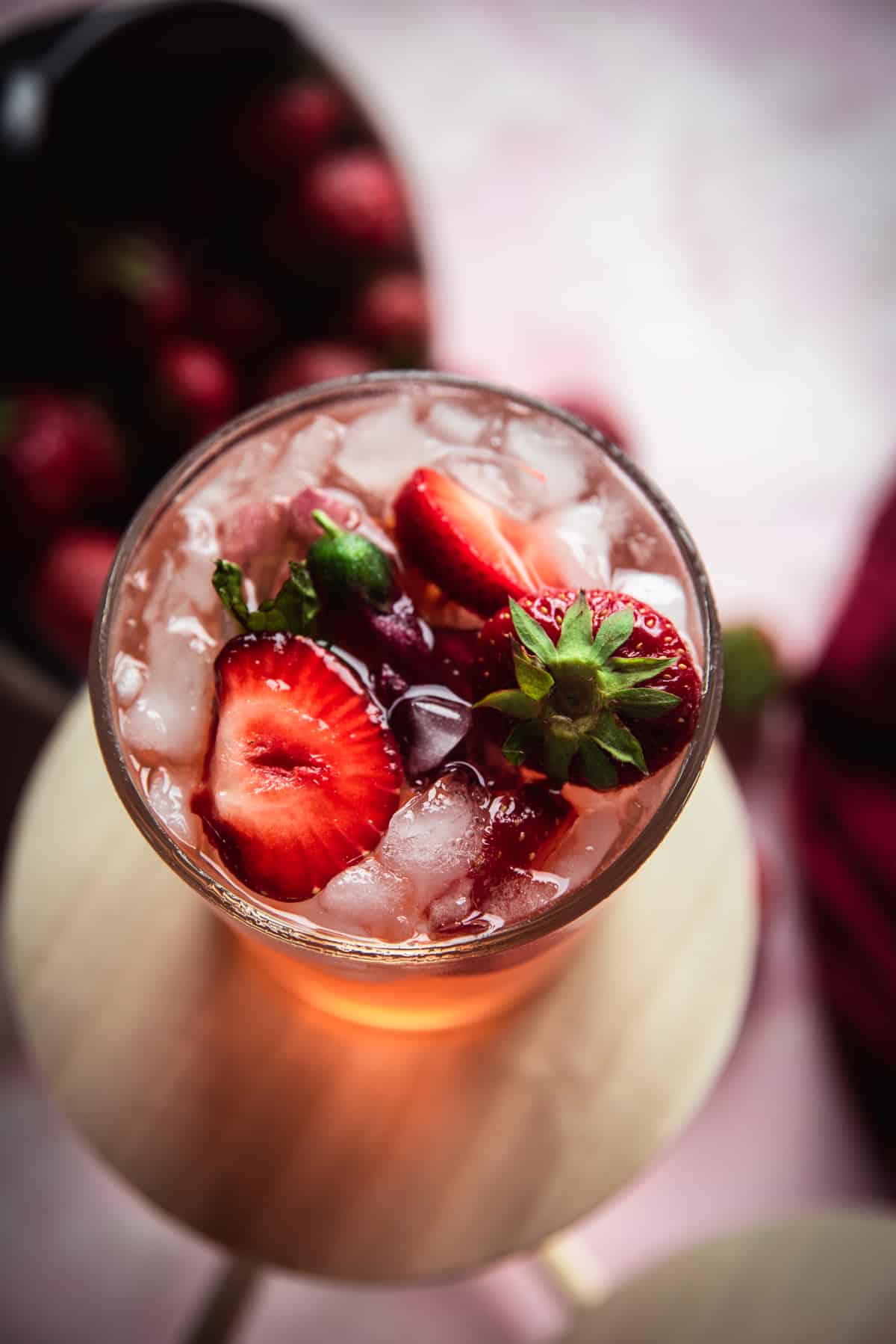 Overhead POV of pink sangria in a glass on top of wooden circle topped with ice and strawberries with light reflecting through it