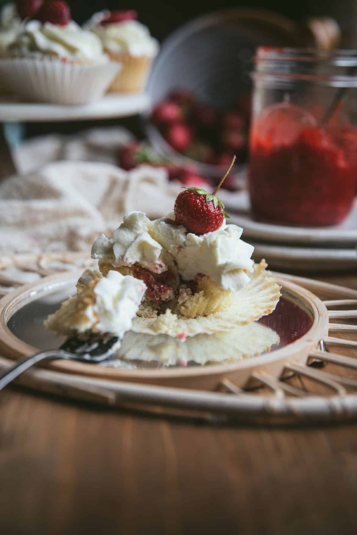 cupcake on a mirrored surface with a forkful take out of it so you can see the strawberry filling with fresh strawberries and more cupcakes in the background