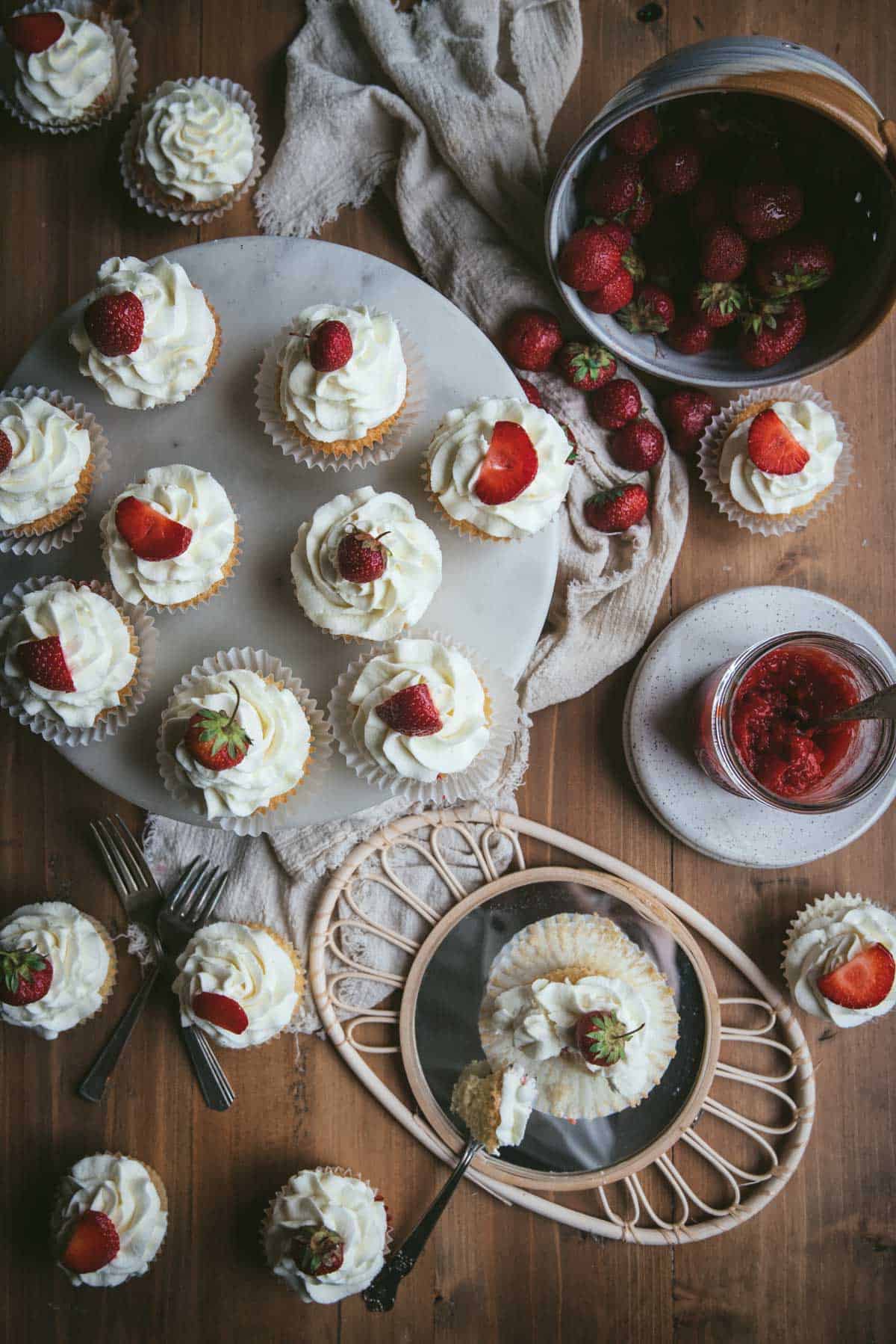 overhead image of 2 dozen cupcakes on top of wooden surface and cupcake stand with fresh strawberries and strawberry filling on the table.