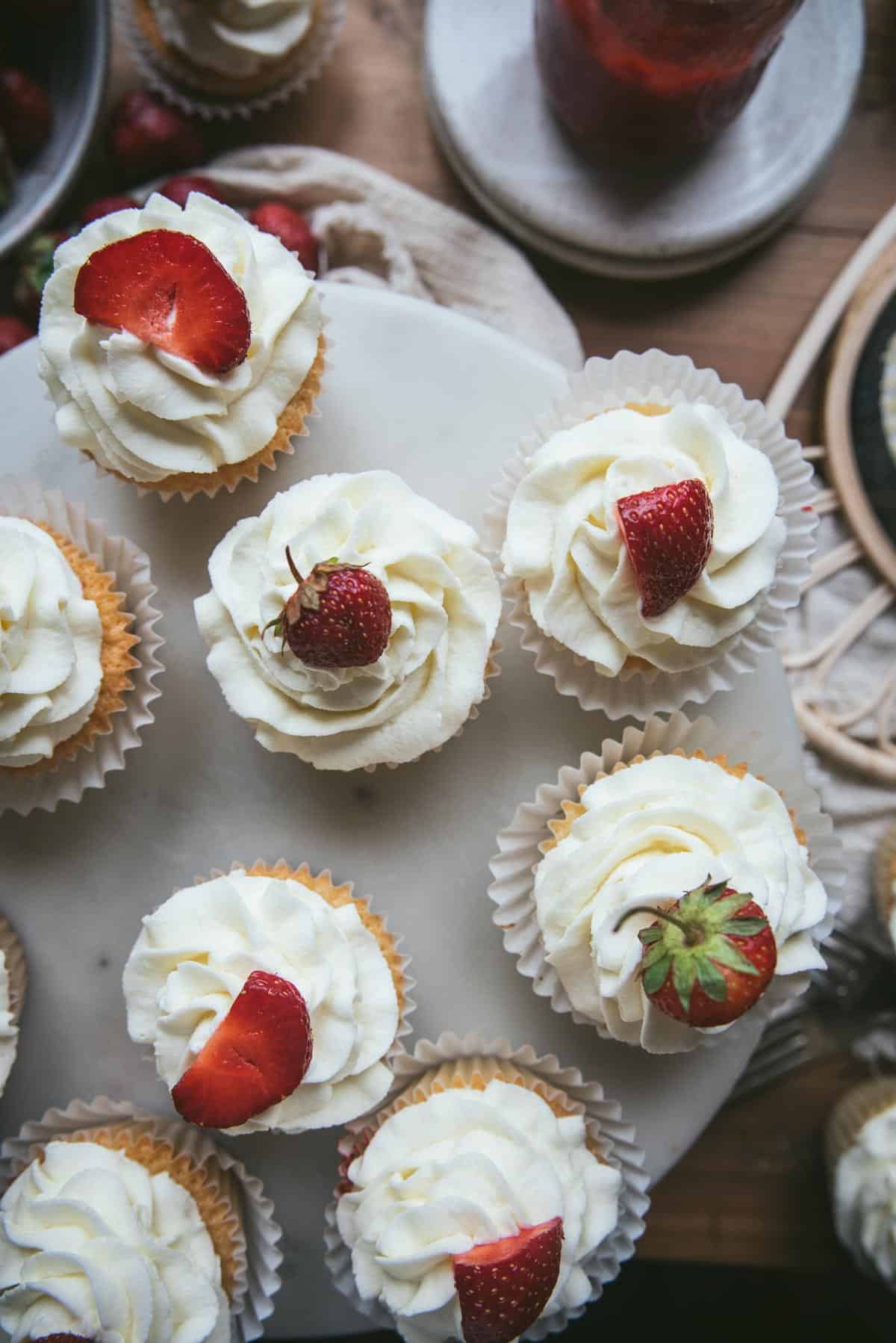 overhead image of whipped cream topped cupcakes with strawberry slices or whole strawberries on garnish.  All the cupcakes are on a cake stand on top of a table.