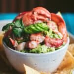 close up of guacamole in a speckled white bowl topped with large chunks and claws of lobster meat, thin slices of jalapeno, and a spring of cilantro.