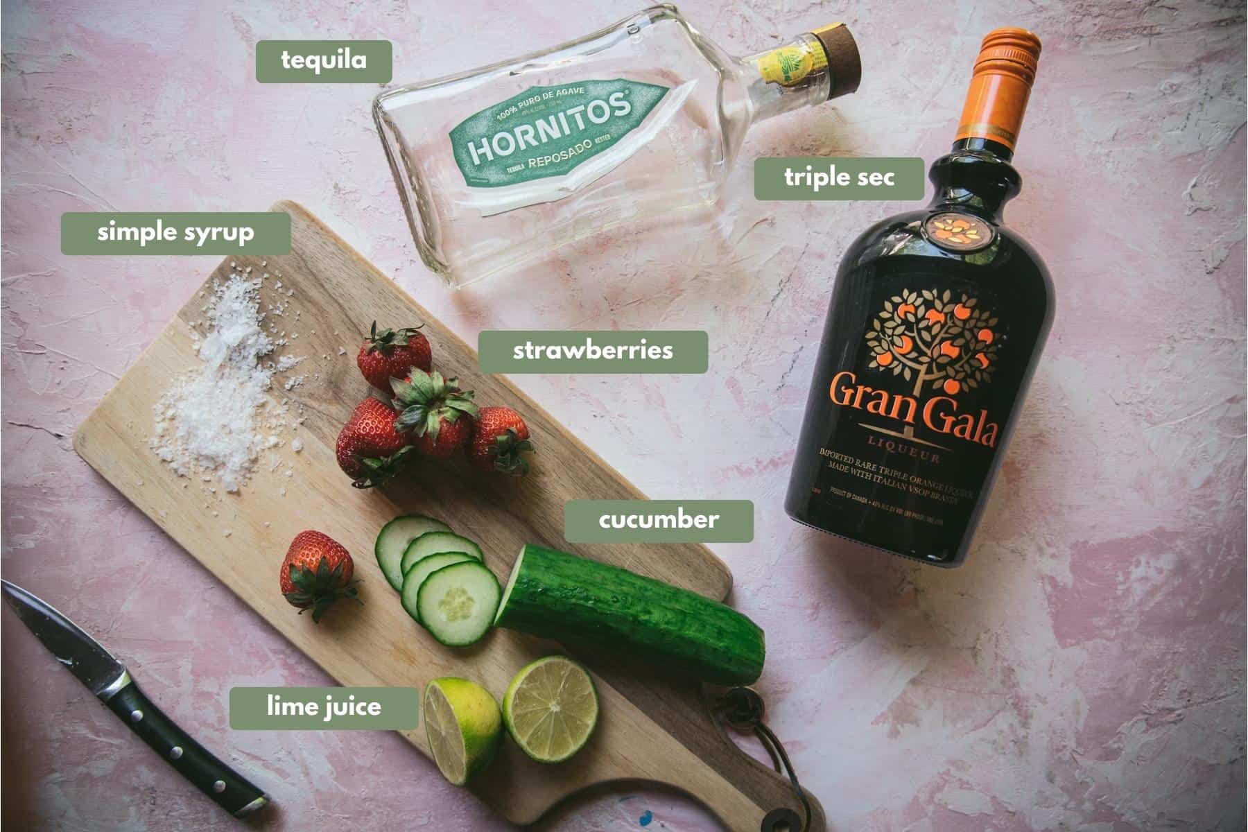 Ingredients for Strawberry Cucumber Margarita. Wooden chopping board with sugar, whole strawberries and cucumber. Next to that is a clear bottle of Hornitos silver tequila and a black botlle Gran Gala.