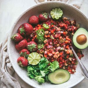 overhead image of large bowl with diced strawberry salsa topped with whole avocado, limes, jalapeno slices, whole strawberries, and cilantro.