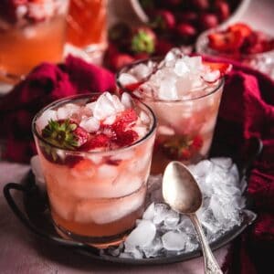 two glasses with ice and pink sangria on a circlular metal serving dish covered in crushed ice and vintage spoon on top. Pitcher of sangria and bowl of strawberries in the background