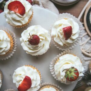 overhead image of whipped cream topped cupcakes with strawberry slices or whole strawberries on garnish. All the cupcakes are on a cake stand on top of a table.
