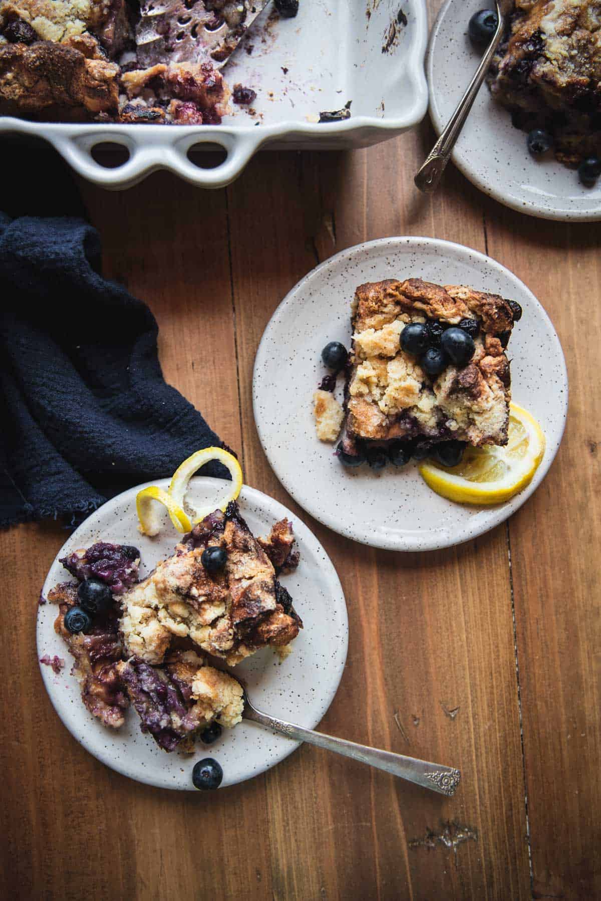 overhead image of two plates with square pieces of french toast casserole baked with blueberries andd topped with fresh blueberries and lemon twists.  Plates are next to the casserole in a baking dish on a blue linen napkin.