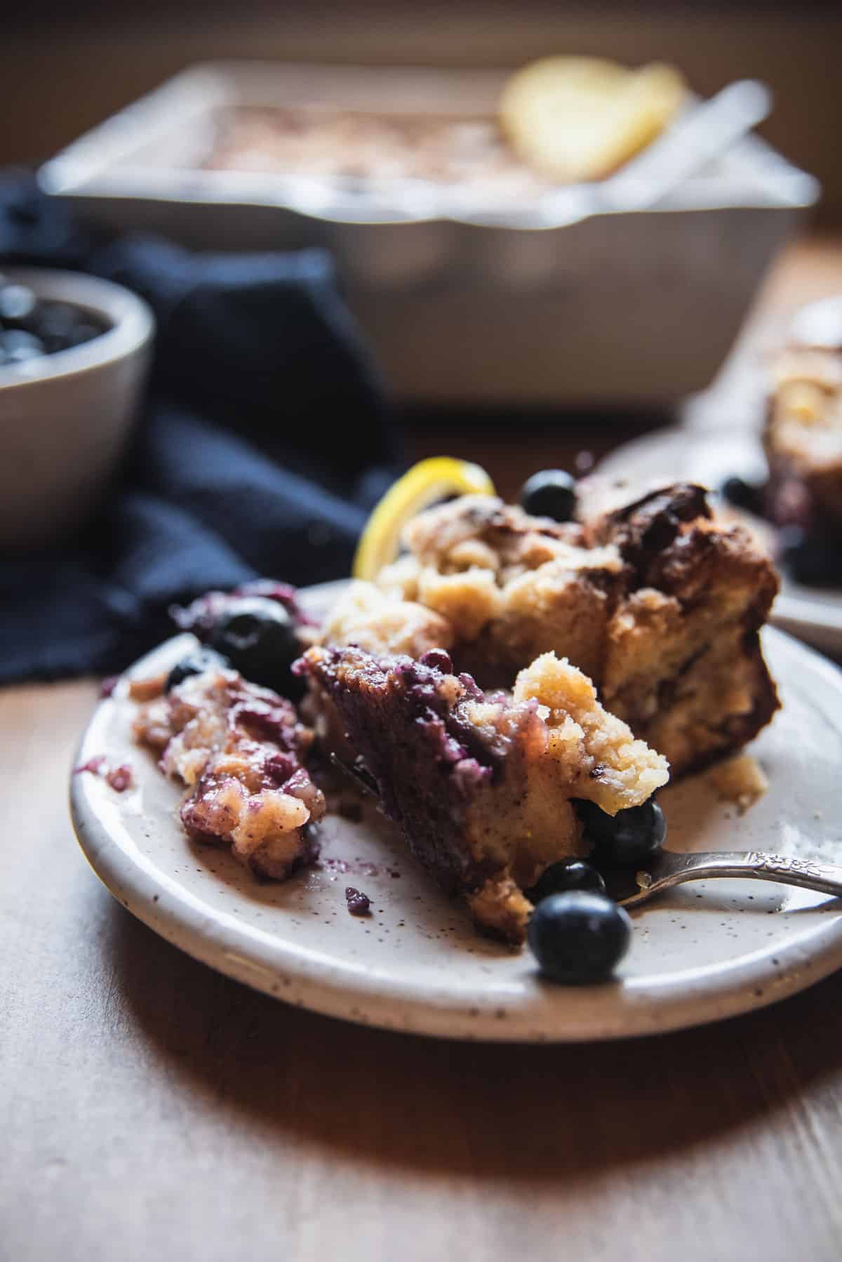 backlit close up photo of fork taking a piece of blueberry french toast casserole on a white speckled plate with baking dishes in the background