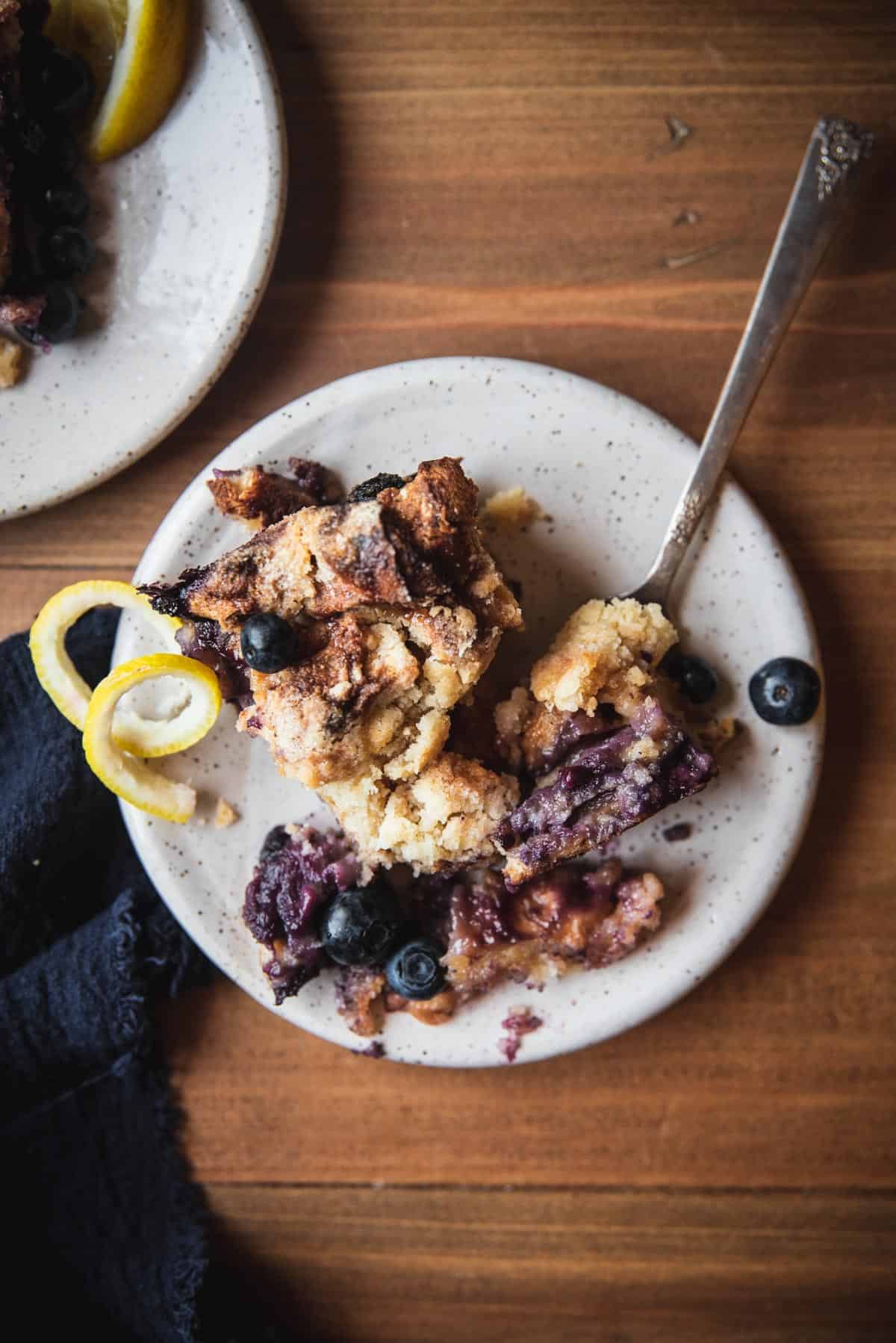 overhead image of baked french toast with blueberries and lemon twist on the plate.  There is an antique fork with a bite on it on the plate.