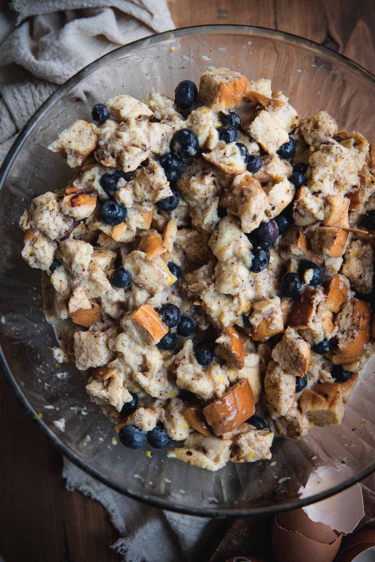 overhead image of bread pieces soaked with french toast ingredients and tossed with fresh blueberries in a large glass mixing bowl.