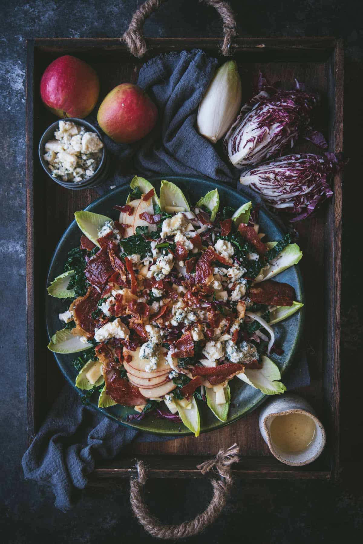 overhead image of salad on a large platter surrounded by whole ingredients.  On the platter there is sliced kale, apples, crispy pieces of prosciutto and crumbled blue cheese on top surrounded by leaves of endive fanned along the outside of the plate.