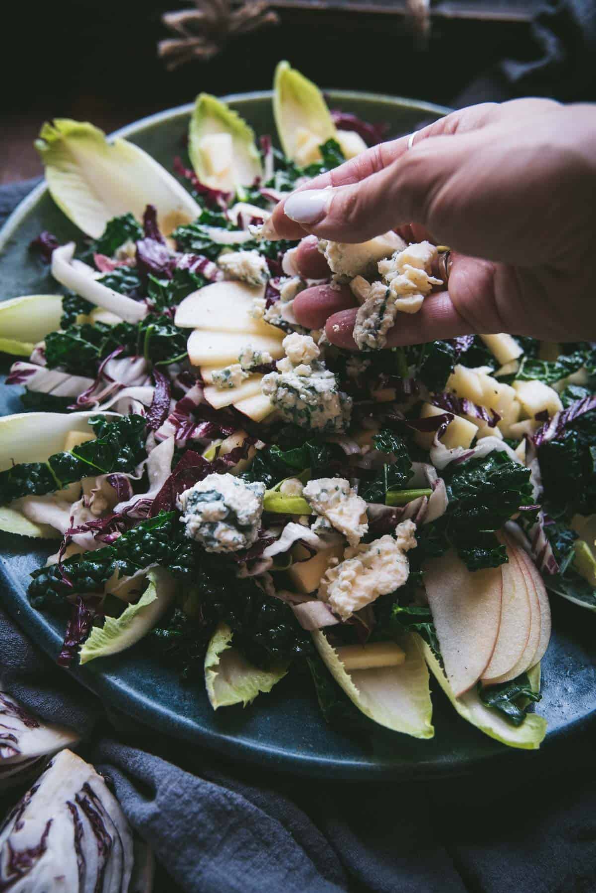 hand crumbling and sprinkling gorgonzola cheese over a salad.  The salad is on a green ceramic plate topped with whole endive leaves, kale, and apples