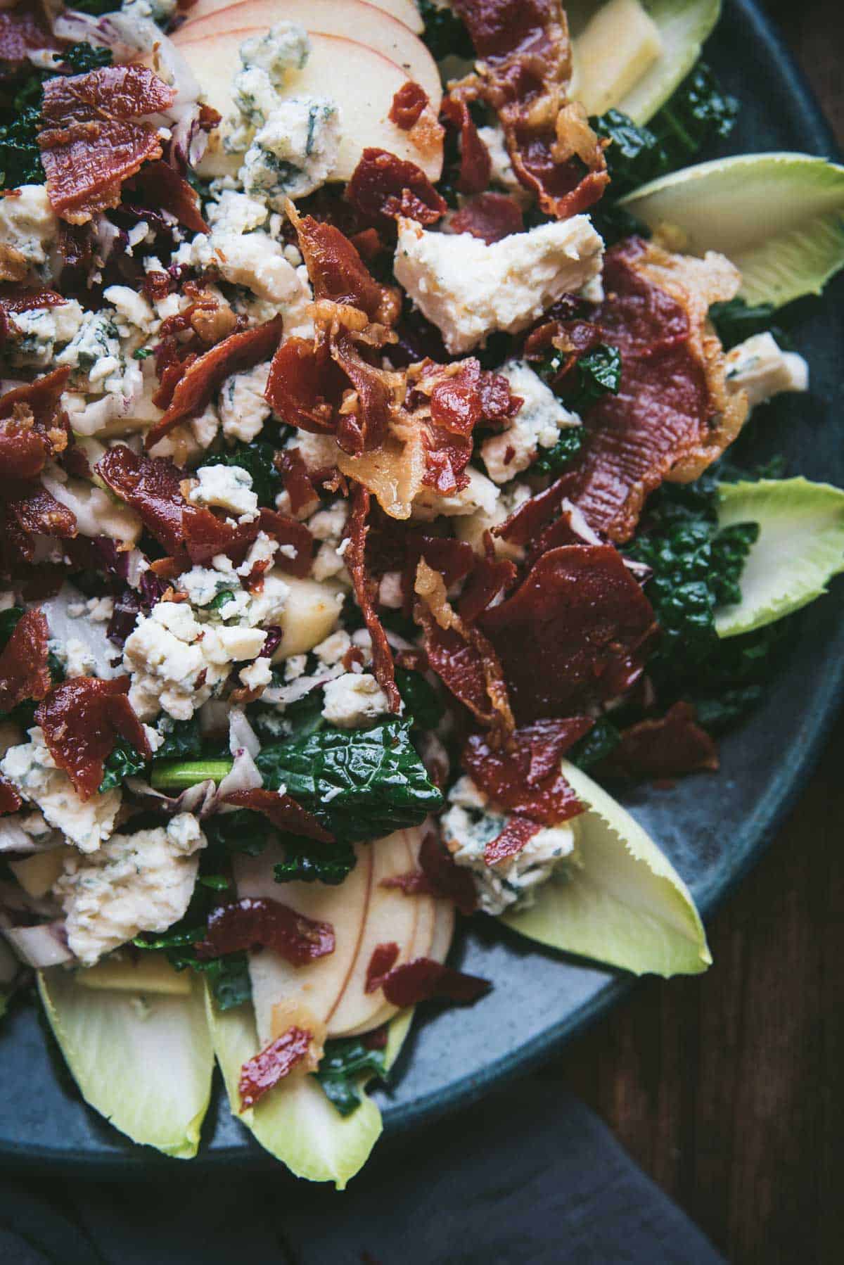 close up of salad on a green ceramic platter with endive, radicchio, kale, gorgonzola cheese, crispy prosciutto on top.