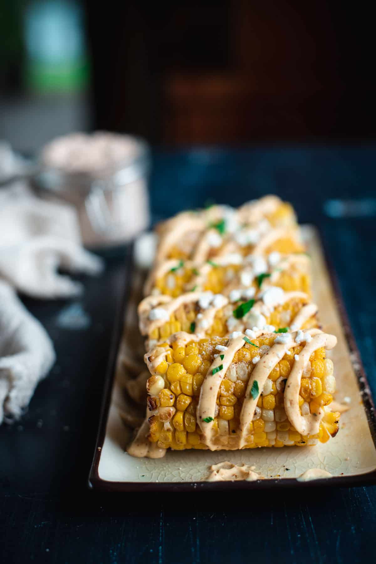 Straight on angle of mexican street corn in a row on a rectangular plate. The corn is topped with a creamy drizzle of sauce, crumbled cheese, and chopped cilantro.