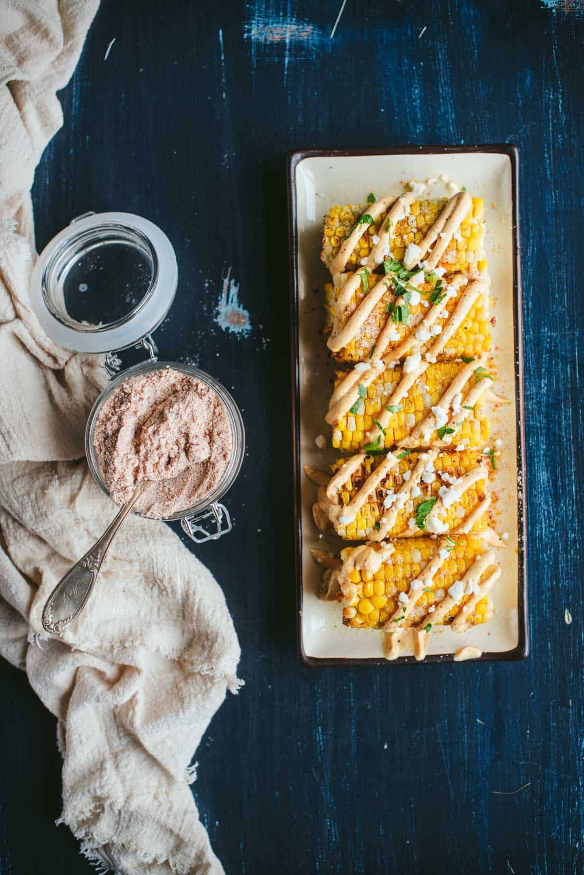 Cooked ears of corn cut in half stacked in a row on a rectangular plate. THe corn is drizzled with a creamy sauce and topped with cheese, cilantro, and seasoning. Plate is next to a jar of elote seasoning.