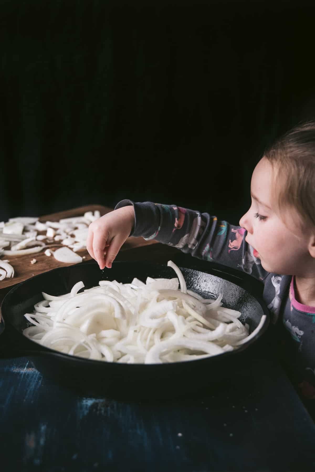 A child is sprinkling salt onto the onions in the cast iron skillet. There is a wooden chopping board with the left over onion on top of.