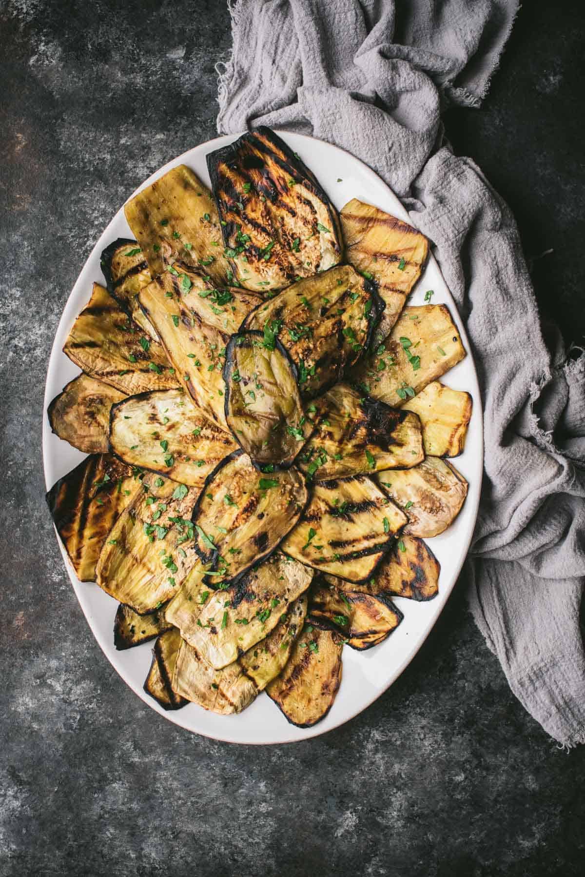 Overhead image of a white platter loaded with perfectly charred grilled eggplant and garnished with herbs with a gray linen napkin beside it.