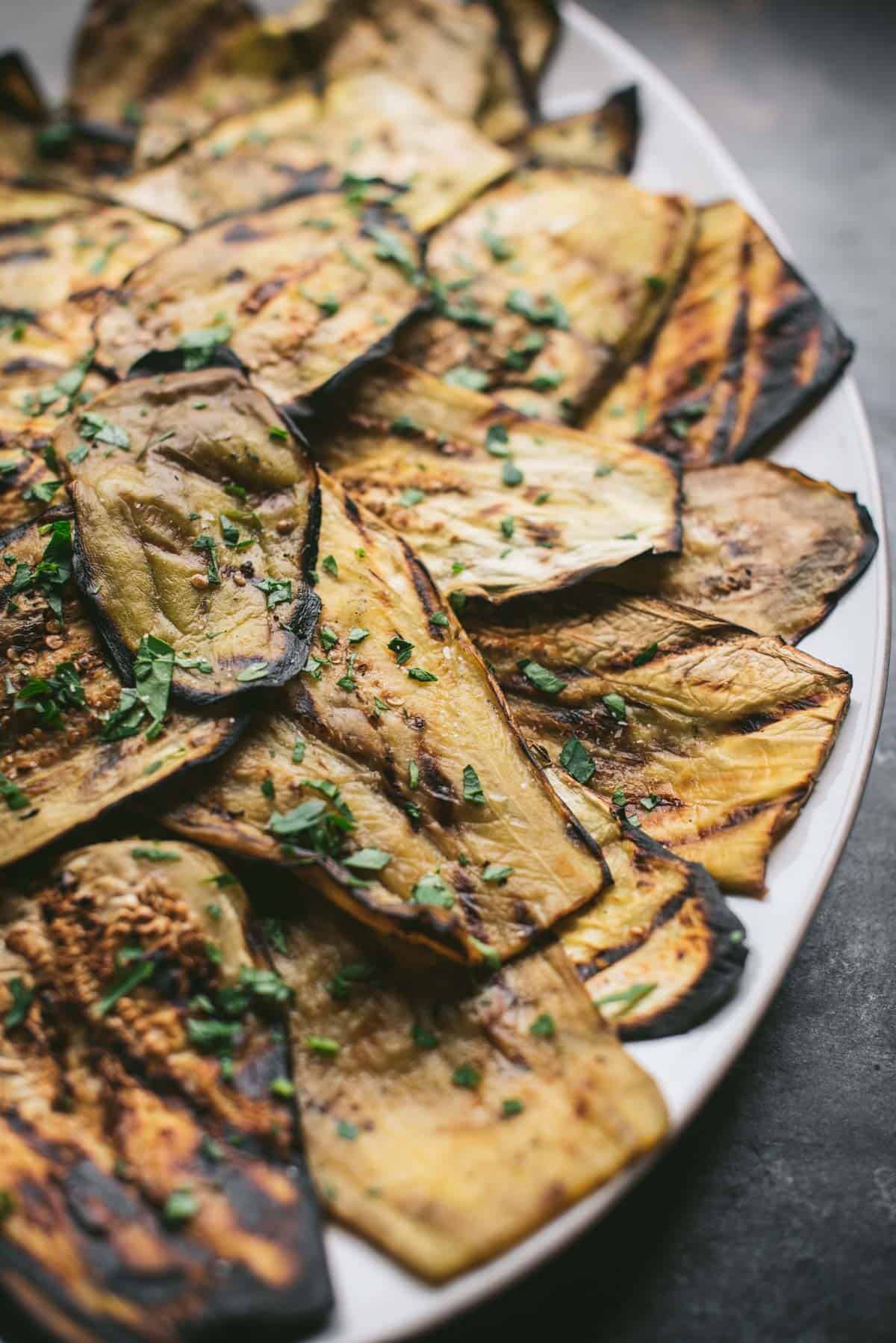 A white platter loaded with perfectly charred grilled eggplant and garnished with herbs.