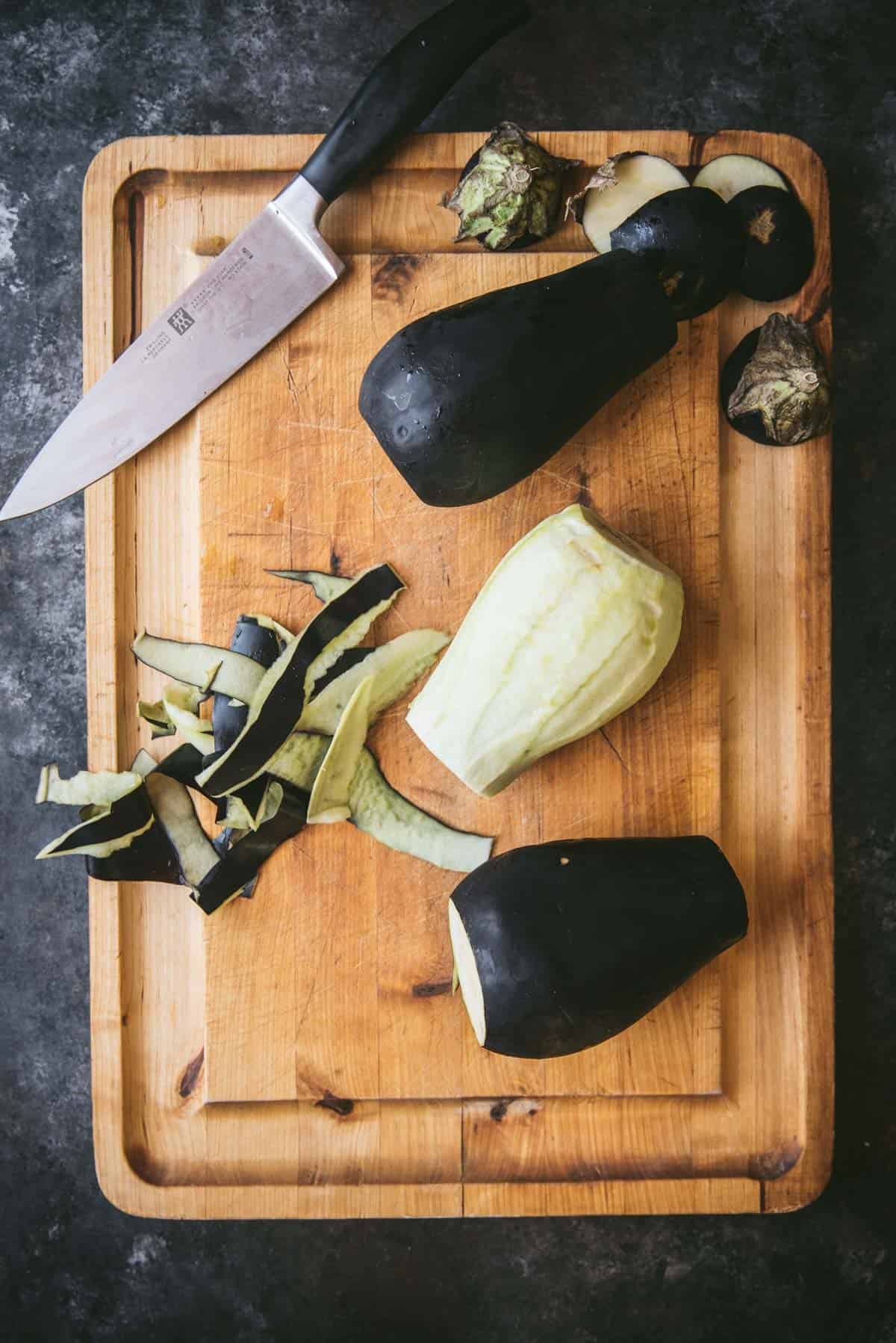 A wooden chopping board with one peeled eggplant, there is 2 more eggplants with the top and bottom chopped off, waiting to be peeled. The knife is laying on the board as is a small pile of eggplant peel.