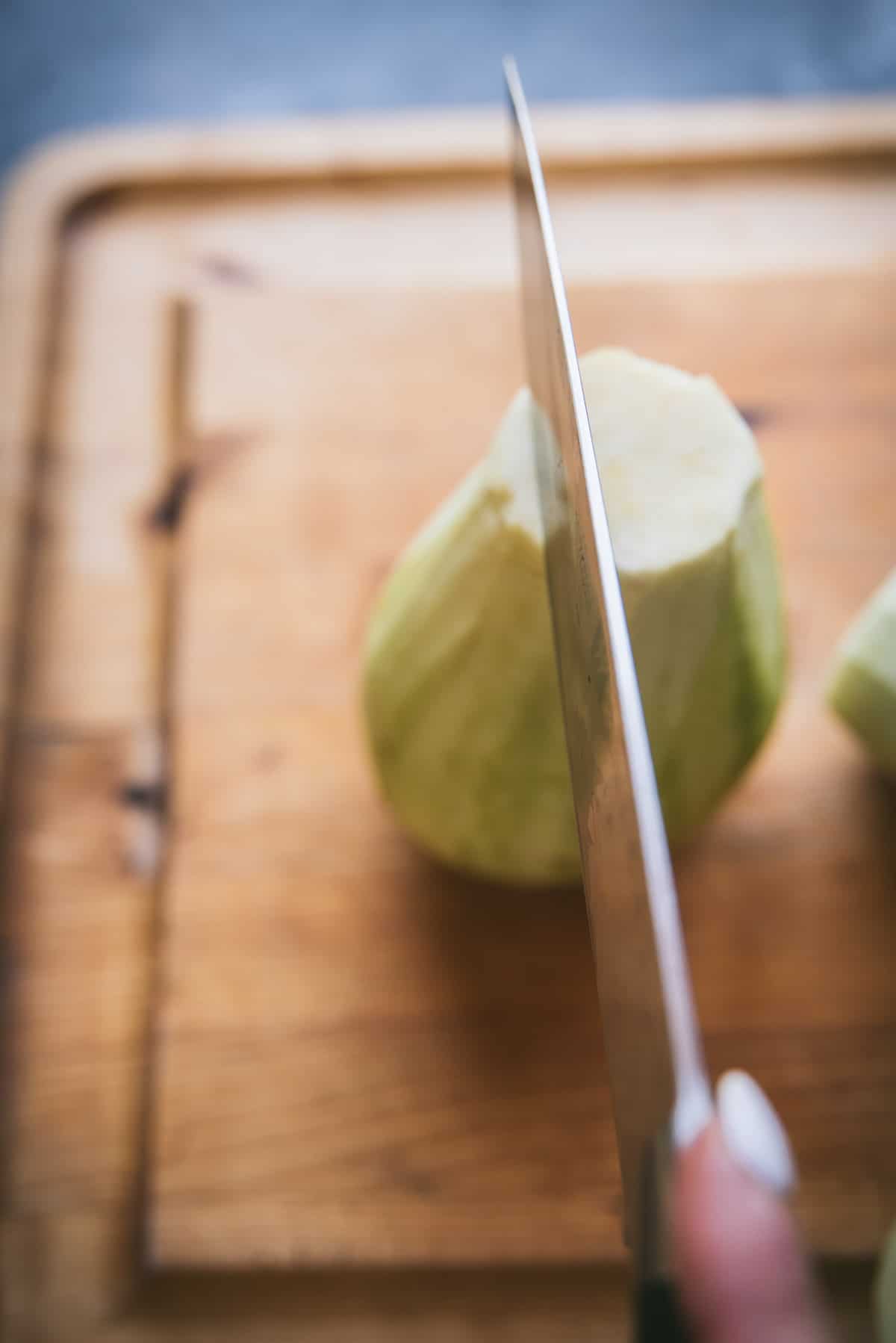 A hand holding a knife is preparing to chop through a peeled eggplant on top of a wooden chopping board.