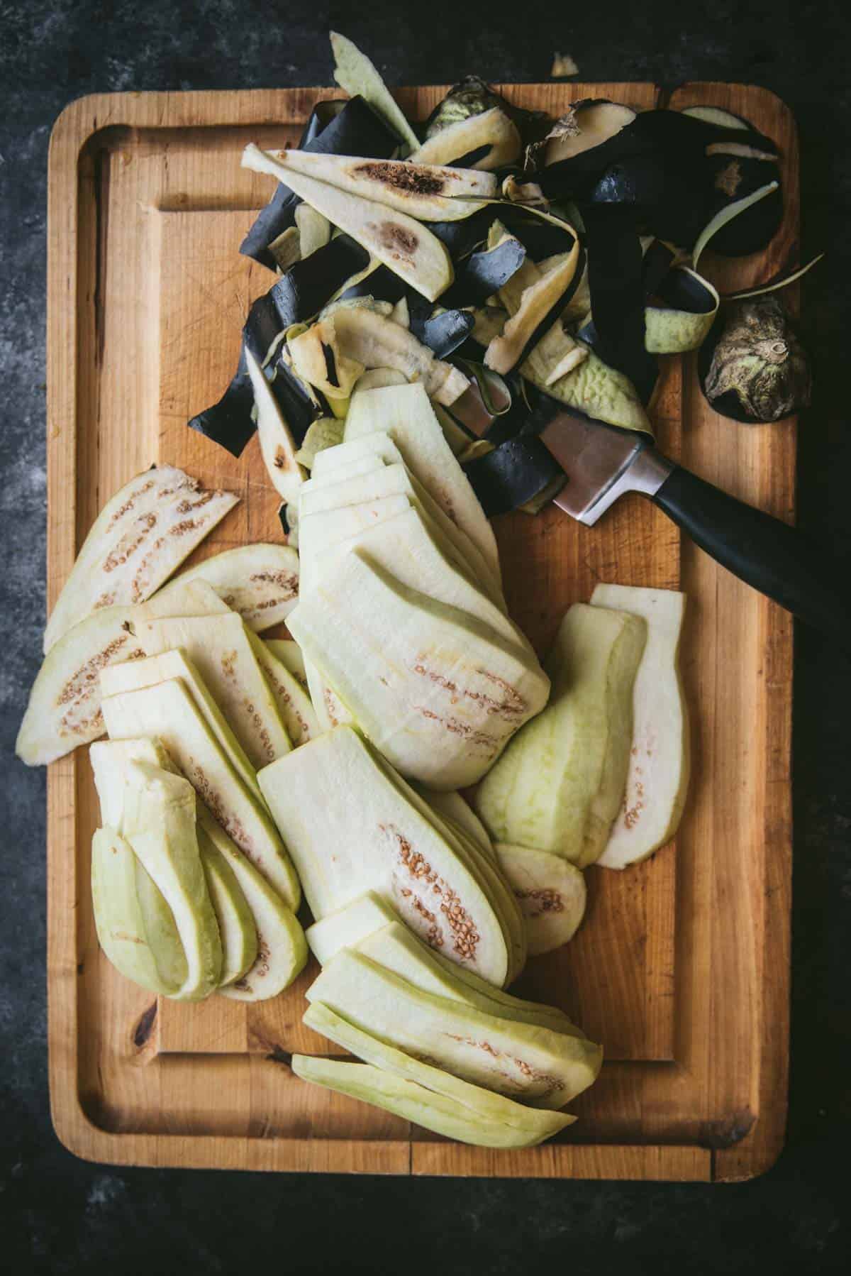 Eggplant is now peeled and sliced into planks atop the wooden chopping board. There is a knife underneath a large pile of eggplant skin waiting to be discarded.