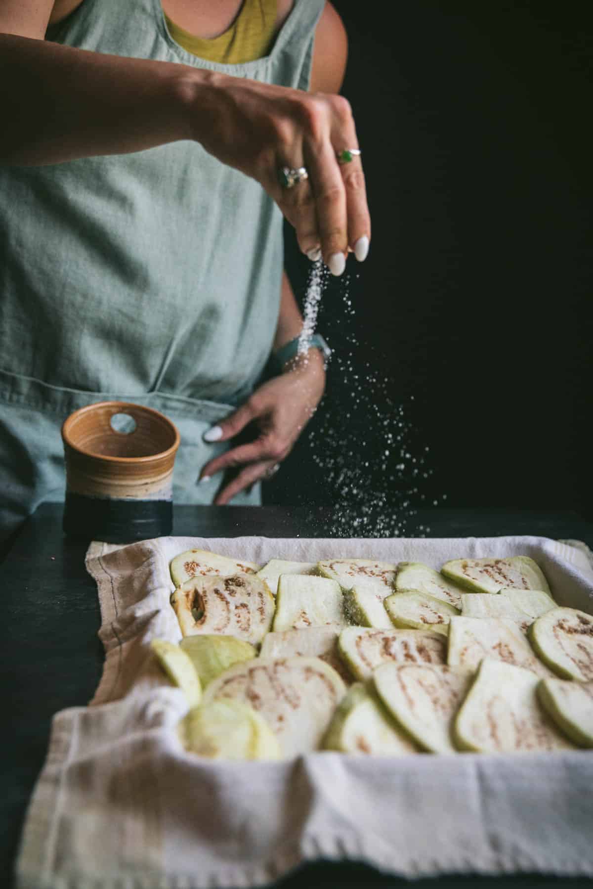 The eggplant is resting in a baking tray on top of a dish towel. All the slices are touching each other on the side and a hand is generously sprinkling salt all over the top of them.