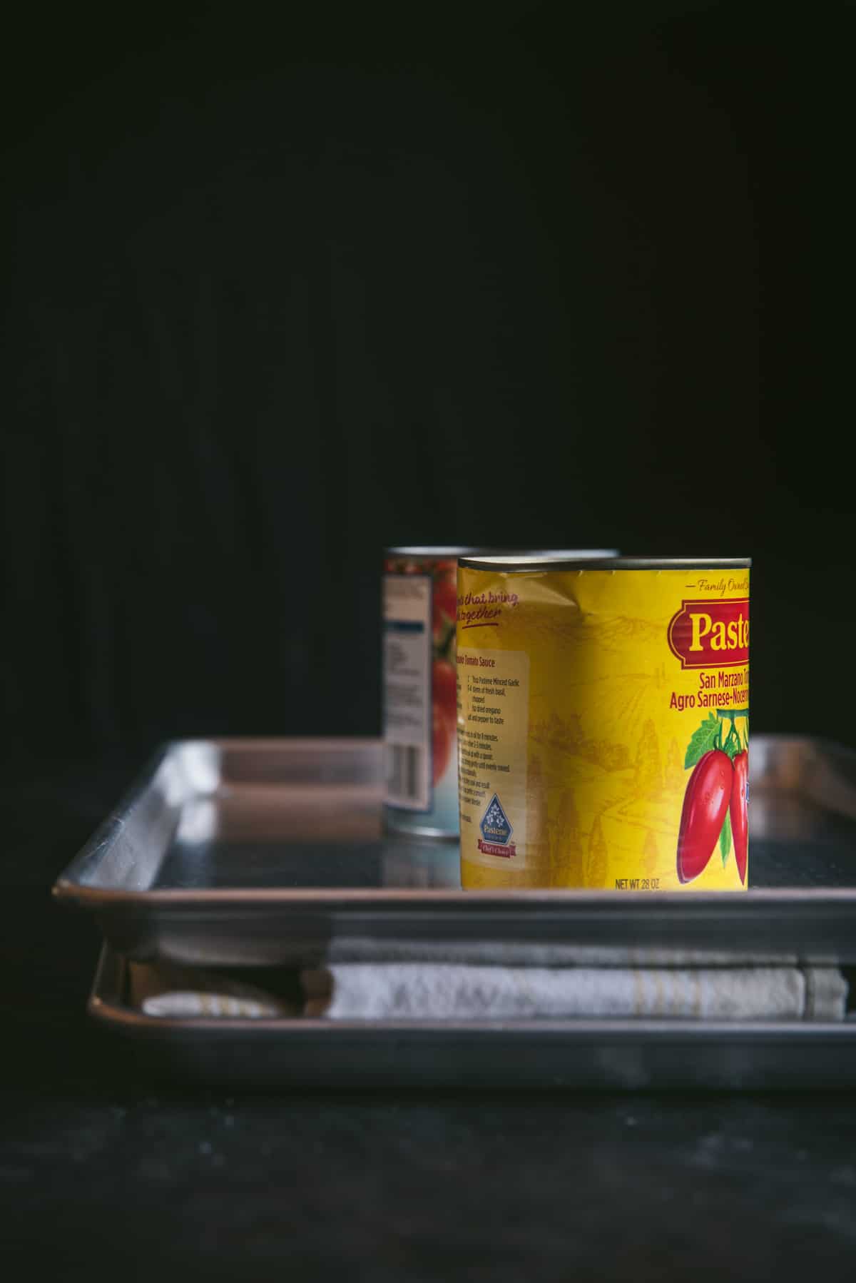 The eggplant is inside of a towel, sandwiched between to baking trays. 2 large cans of tomatoes have been placed on top to press all of the moisture and liquid out of the eggplant.